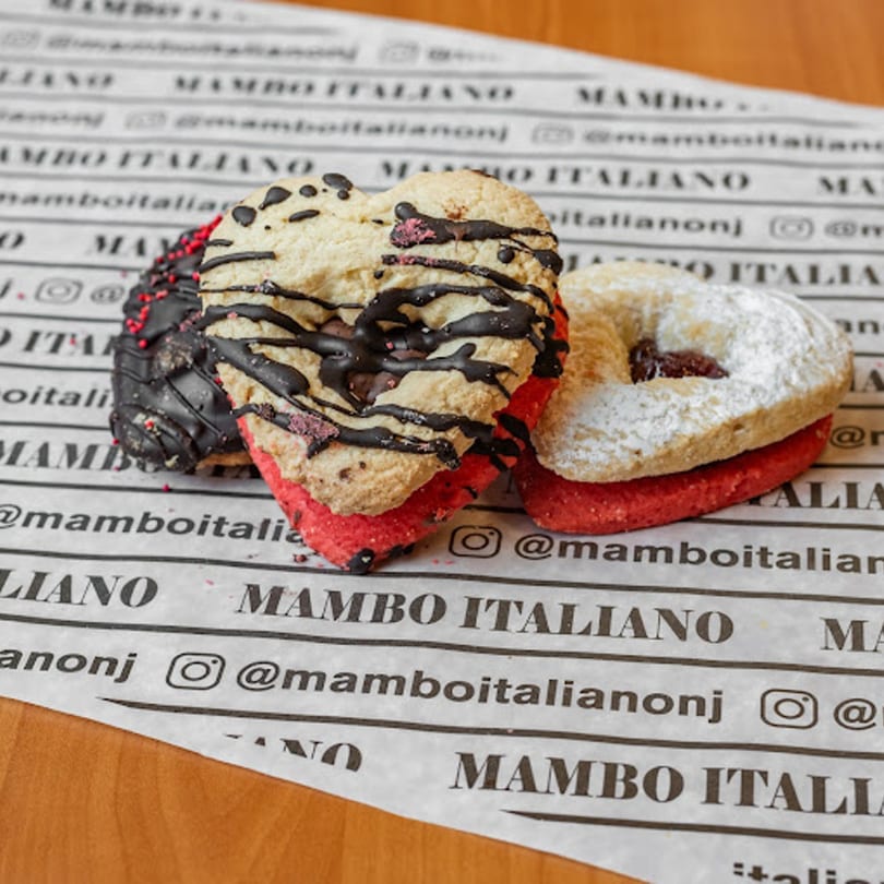 Three heart-shaped Italian cookies, two with chocolate drizzle and one with powdered sugar, on branded paper.