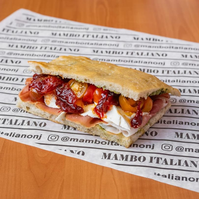 A Mambo Italiano focaccia sandwich with prosciutto, mozzarella, and roasted tomatoes on branded paper.